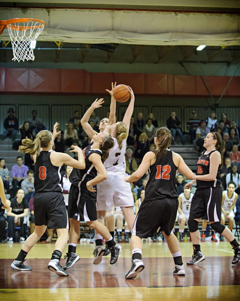 A basketball player in white jumps to shoot while surrounded by opposing players in black jerseys during a game.