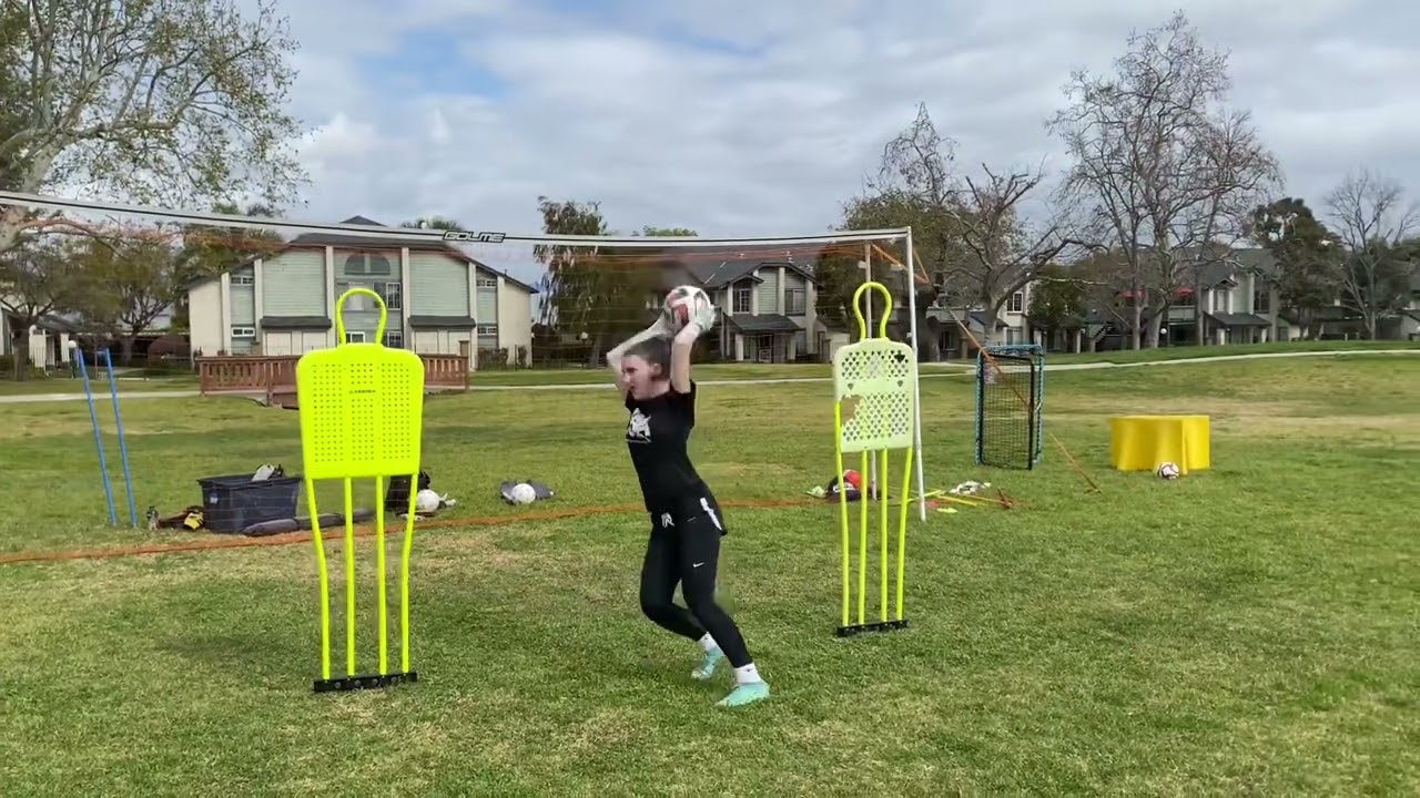 Person practicing a soccer throw-in between two yellow training mannequins on a grass field with houses in the background.