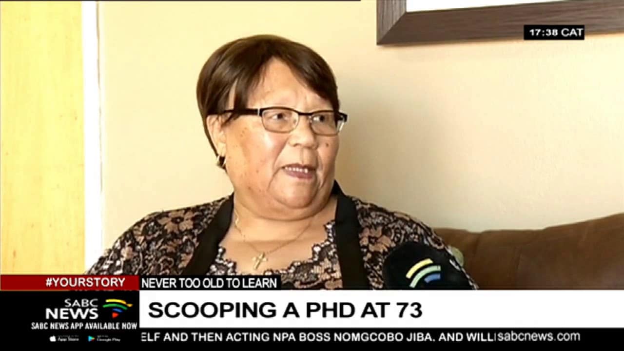 An older woman with glasses and a patterned blouse sits indoors, smiling. The text on screen reads: "SCOOPING A PHD AT 73". SABC News logo and timestamp in corners.