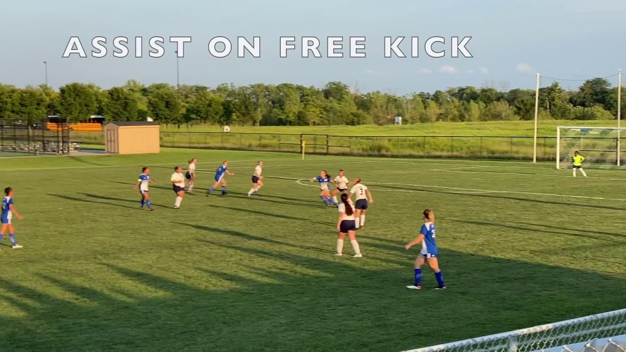 Soccer game in progress on a green field with players in blue and white jerseys. Text reads "Assist on Free Kick" at the top of the image.