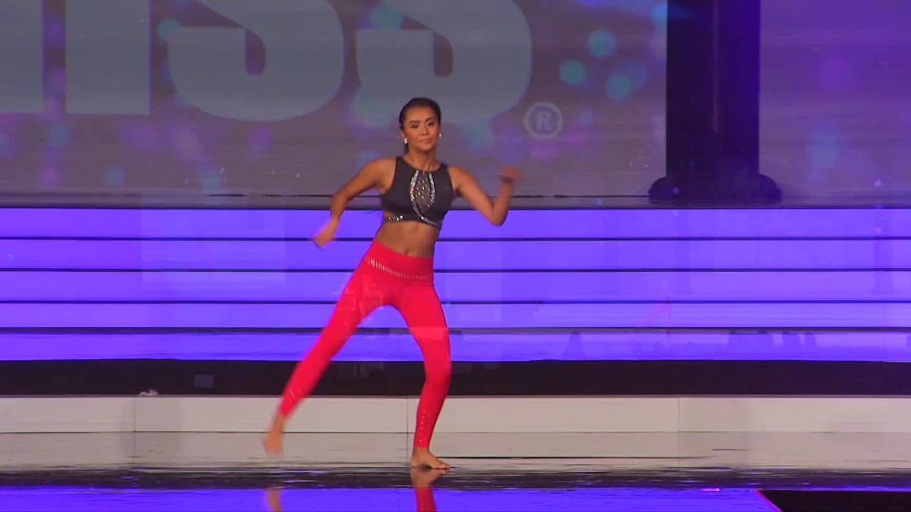 A dancer performs energetically on stage wearing a black top and red pants against a backdrop with blue and purple lighting.