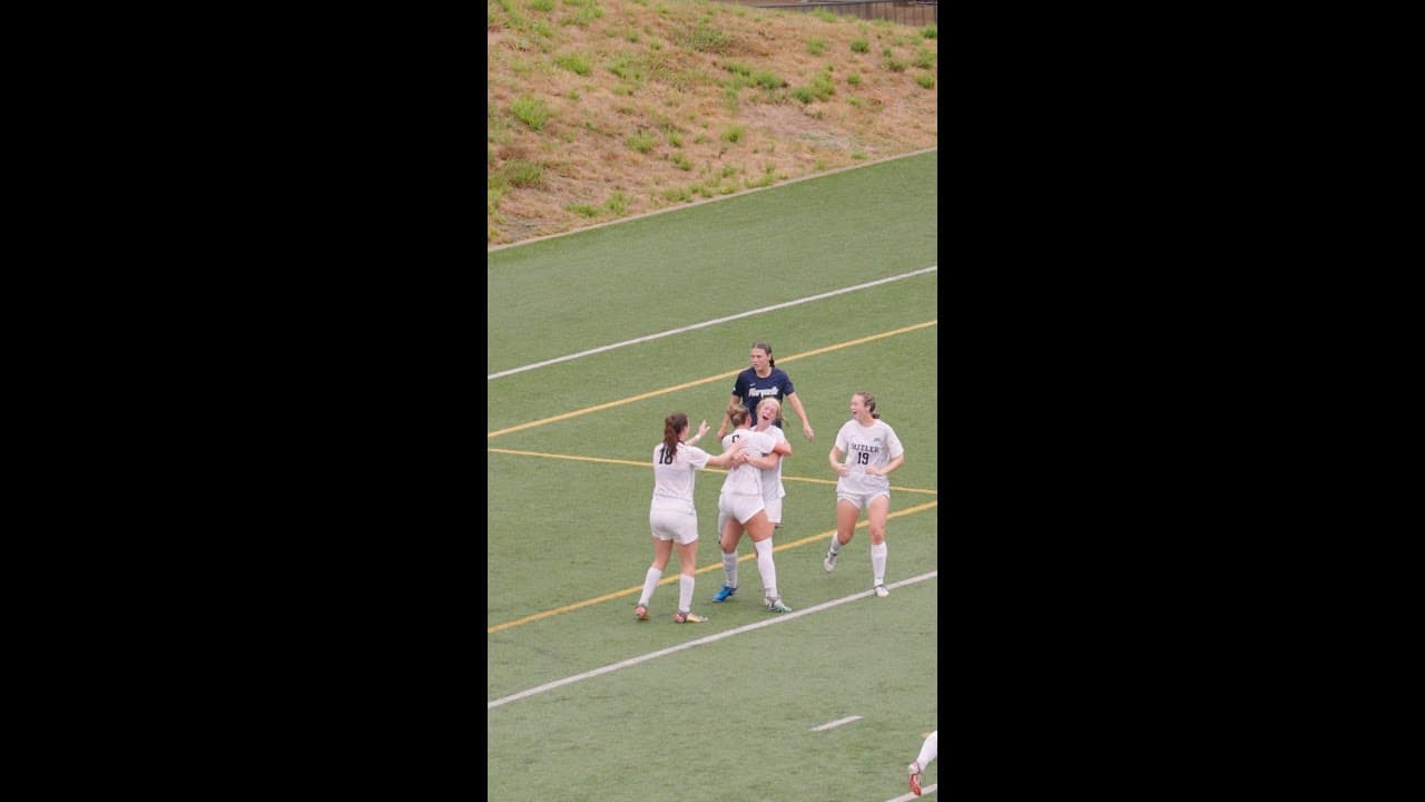 Soccer players in white uniforms celebrate on a field as one player in a navy uniform walks nearby.