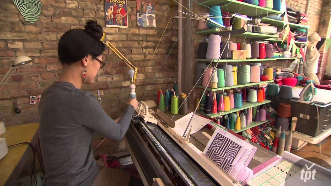 Person using a knitting machine in a workshop with shelves of colorful yarn cones and a brick wall.