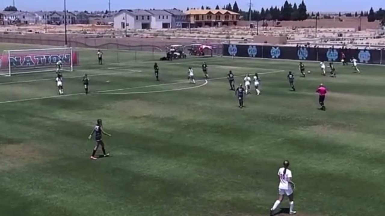 Soccer match in progress on a grassy field, players in dark and white uniforms, with houses and construction in the background.