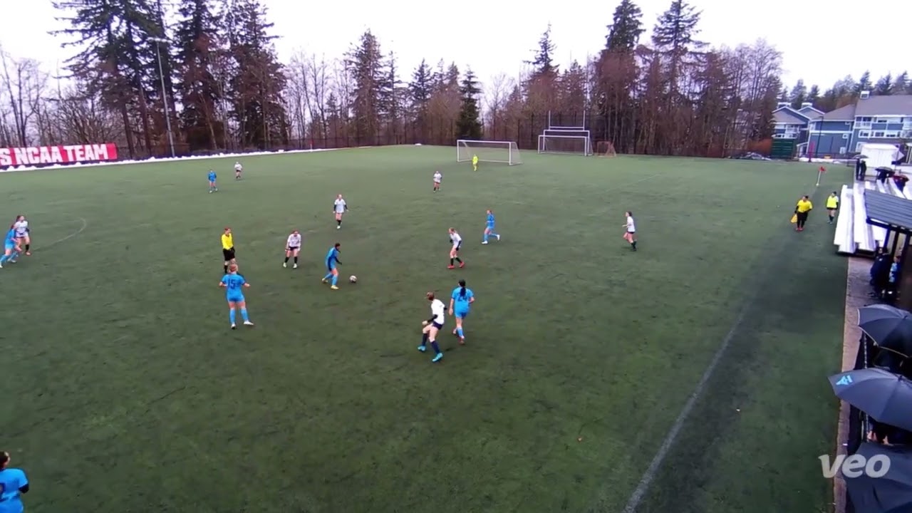 Aerial view of a soccer game in progress on a green field, with players in blue and white uniforms. Trees and a banner with "NCAA TEAM" are visible in the background.