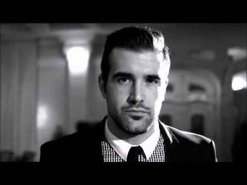 Man in a suit with a checkered shirt and a tie, standing in a dimly lit room with blurred pillars in the background. Black and white image.