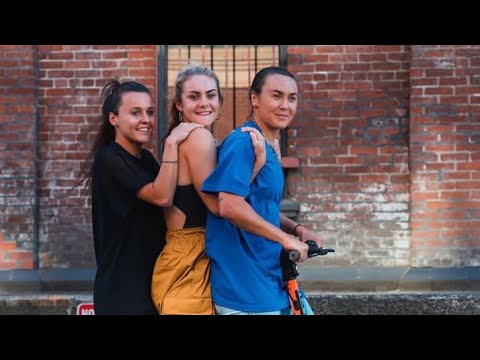 Three people ride together on a scooter against a brick wall backdrop, smiling at the camera.