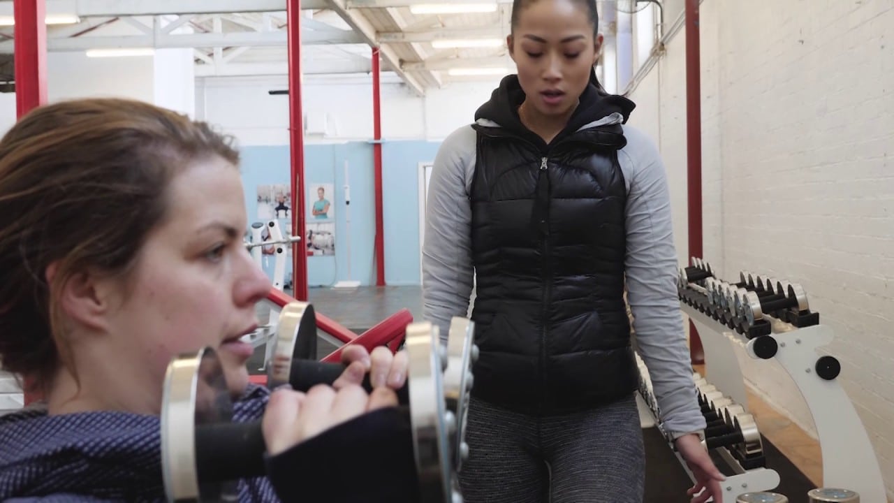 Two women are in a gym. One is seated, holding a dumbbell, performing an exercise. The other is standing nearby, appearing to instruct or observe. Fitness equipment is visible in the background.