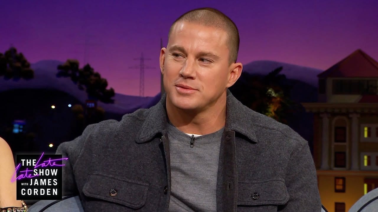 A person sits on "The Late Late Show with James Corden" set, wearing a gray shirt and jacket, against a backdrop featuring buildings and mountains.