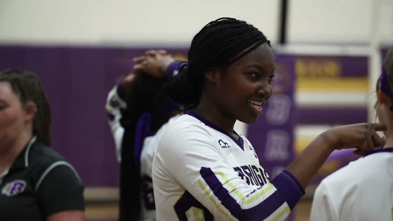 A volleyball player in a white and purple jersey gestures while talking with teammates.