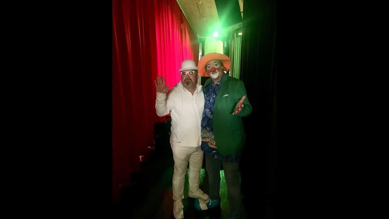 Two men in vibrant costumes and hats pose together under stage lights, one in white and the other in multicolored attire, against a backdrop of red curtains.