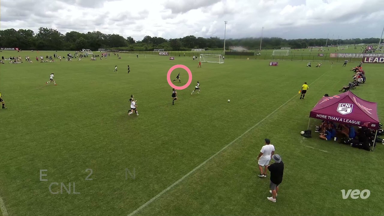 Wide-angle view of a soccer match on a grass field, with players and a referee in action. Spectators and a tent are visible on the sidelines.