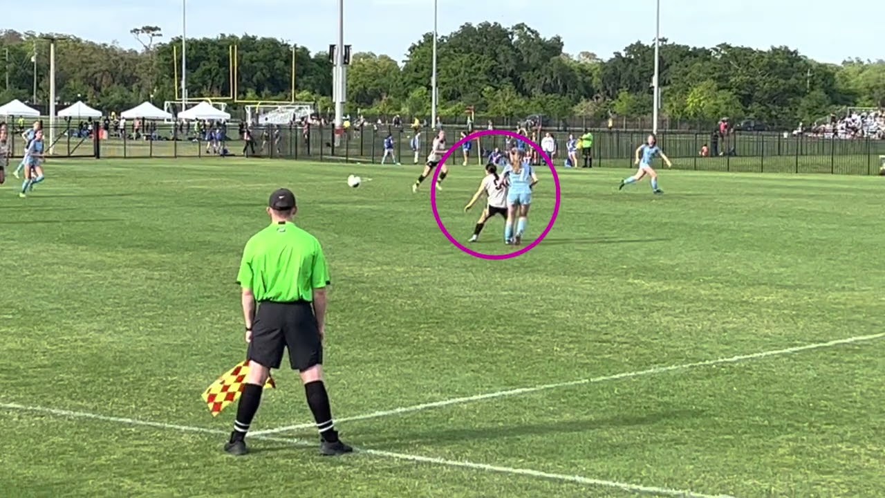 Soccer match with a referee in the foreground, a player crossing the ball, and two players highlighted in a pink circle.