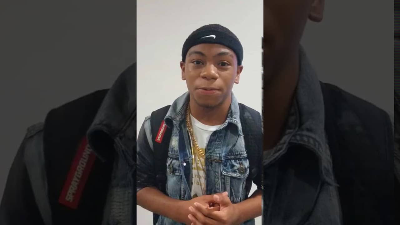 A person wearing a denim jacket, black headband, and backpack stands indoors facing the camera.