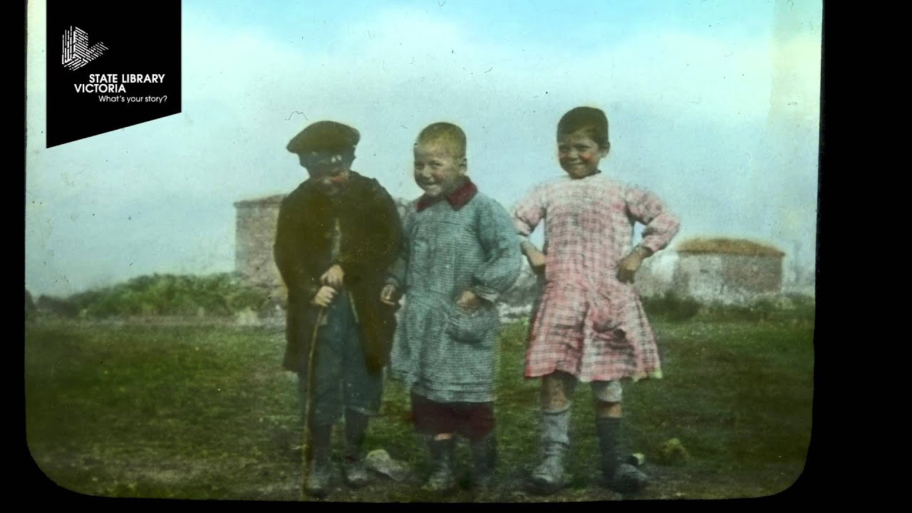 Three children stand together outdoors. One holds a stick, and each is wearing different patterned clothing. There's a building and grassy field in the background.