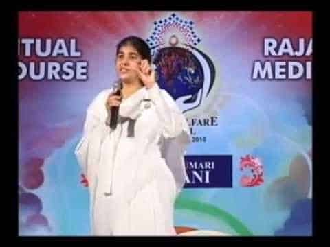 A person in white attire speaks on stage with a microphone, standing in front of a colorful backdrop displaying text about a spiritual course and Rajayoga meditation.