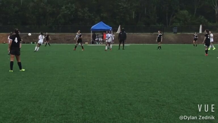 Soccer players on a green field, a player wearing a white uniform kicking the ball. Others in black uniforms observe. A blue tent in the background with people nearby.