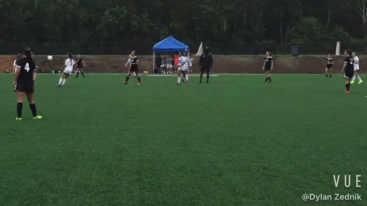 Soccer players on a green field, a player wearing a white uniform kicking the ball. Others in black uniforms observe. A blue tent in the background with people nearby.