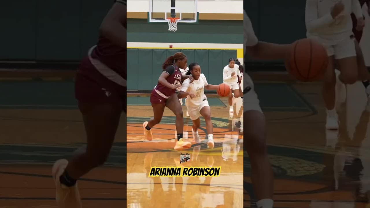 Two basketball players in motion on a court; one in a maroon jersey and the other in a white jersey dribbling the ball.