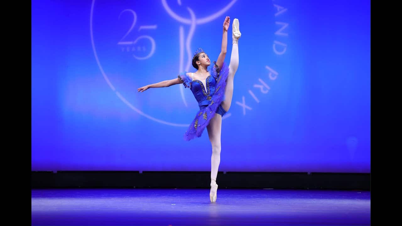 Ballet dancer in a blue tutu balances en pointe with one leg extended high, in front of a blue backdrop with an emblem.