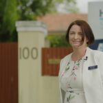 A woman in a white blazer stands in front of a building with a "SOLD" sign and the number 100 on a pillar, smiling at the camera.