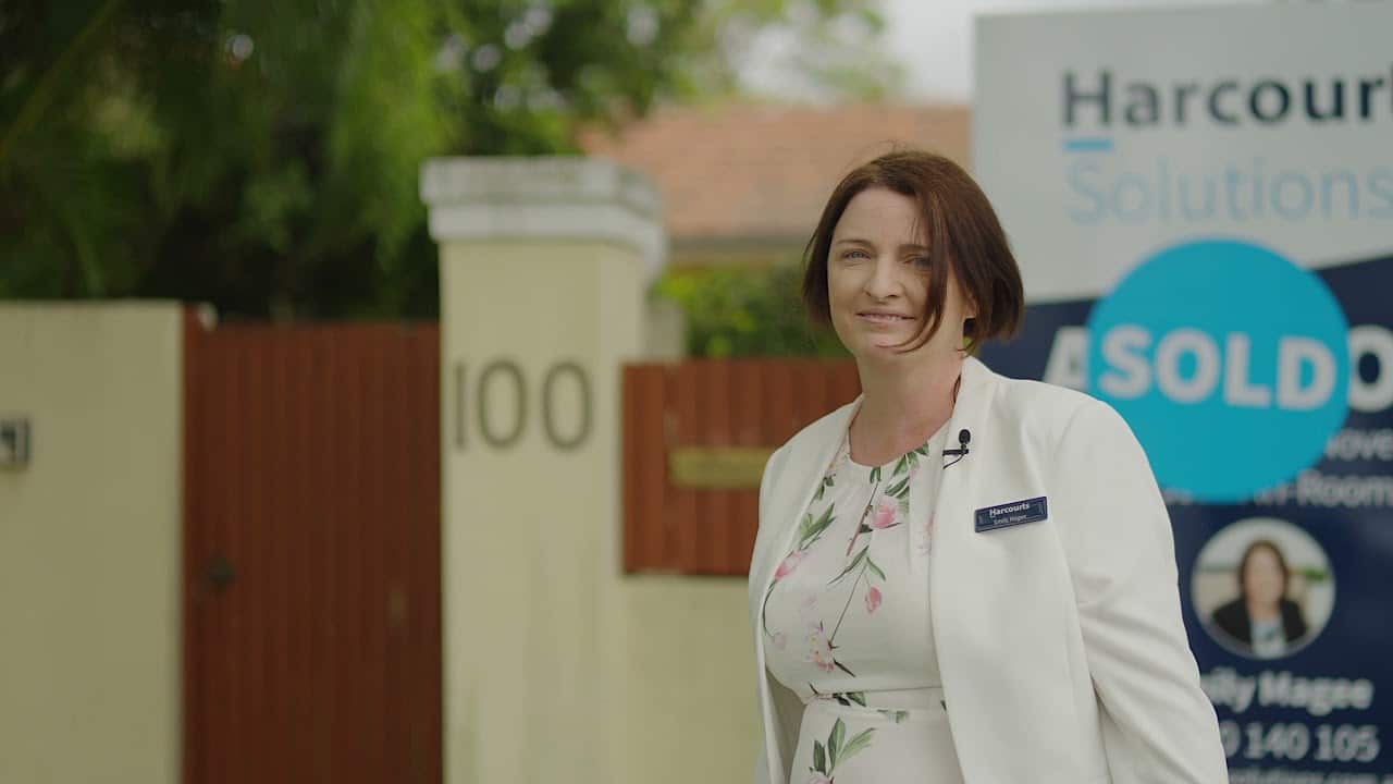 A woman in a white blazer stands in front of a building with a "SOLD" sign and the number 100 on a pillar, smiling at the camera.