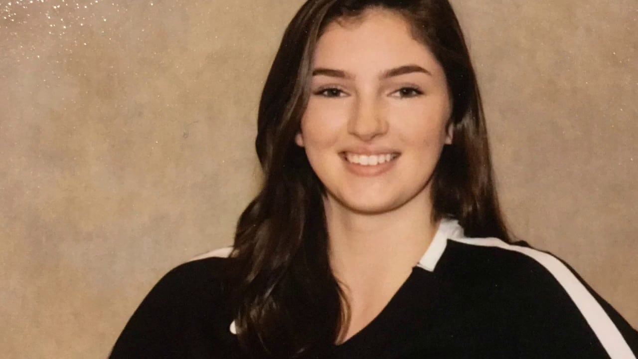 A person with long brown hair is smiling, wearing a black top with white trim, against a plain background.