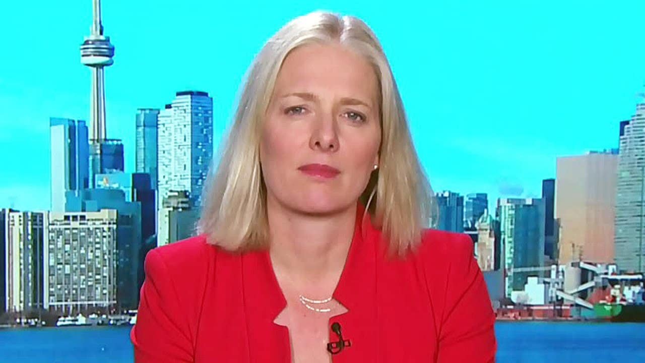 A person in a red blazer sits with a cityscape and tower in the background under a clear blue sky.