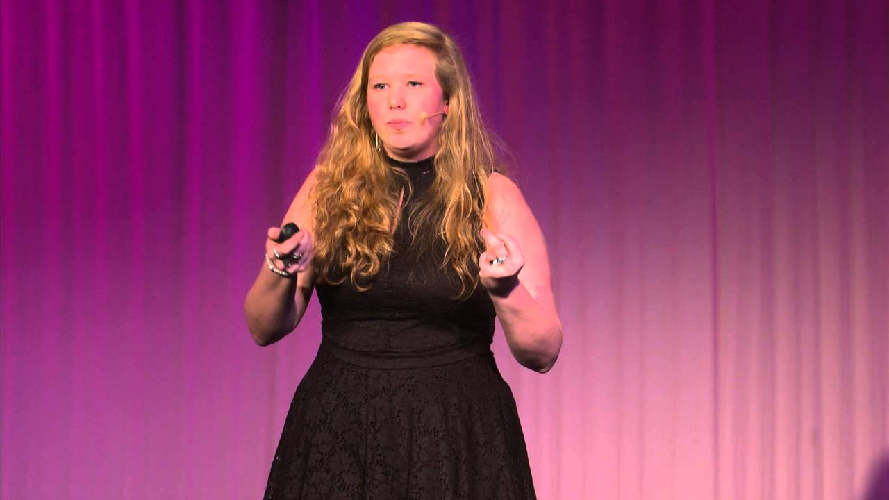 A woman with long blond hair stands on stage with a microphone, wearing a black dress, against a purple background. She is gesturing with her hands while speaking.