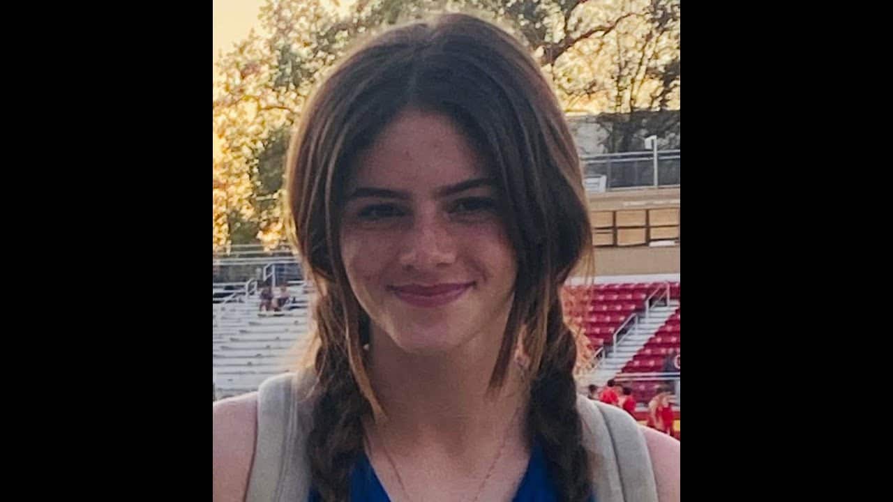 A person with long hair in braids smiles outdoors, standing in front of bleachers with trees in the background.