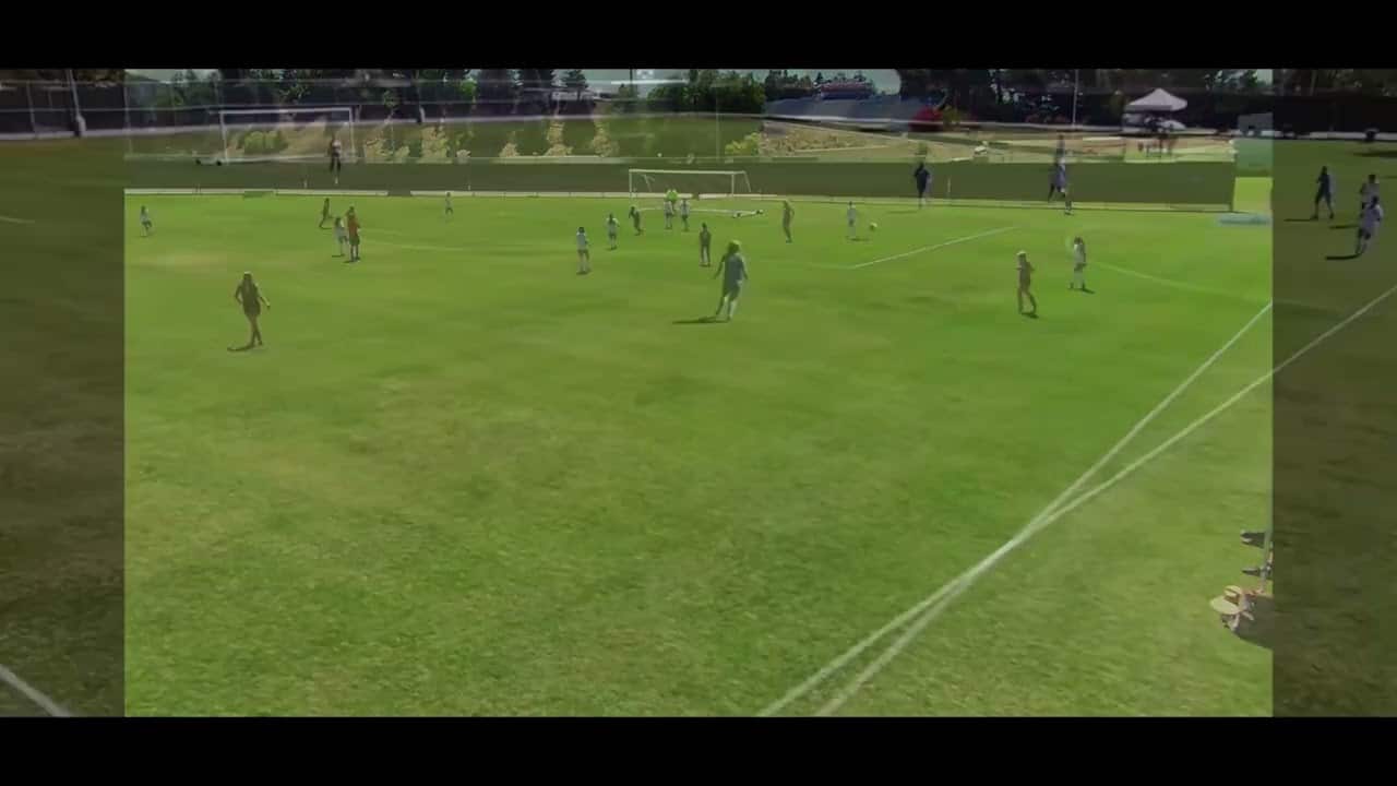 Aerial view of a youth soccer game on a grassy field with players spread across the pitch. Spectators are visible on the sidelines.