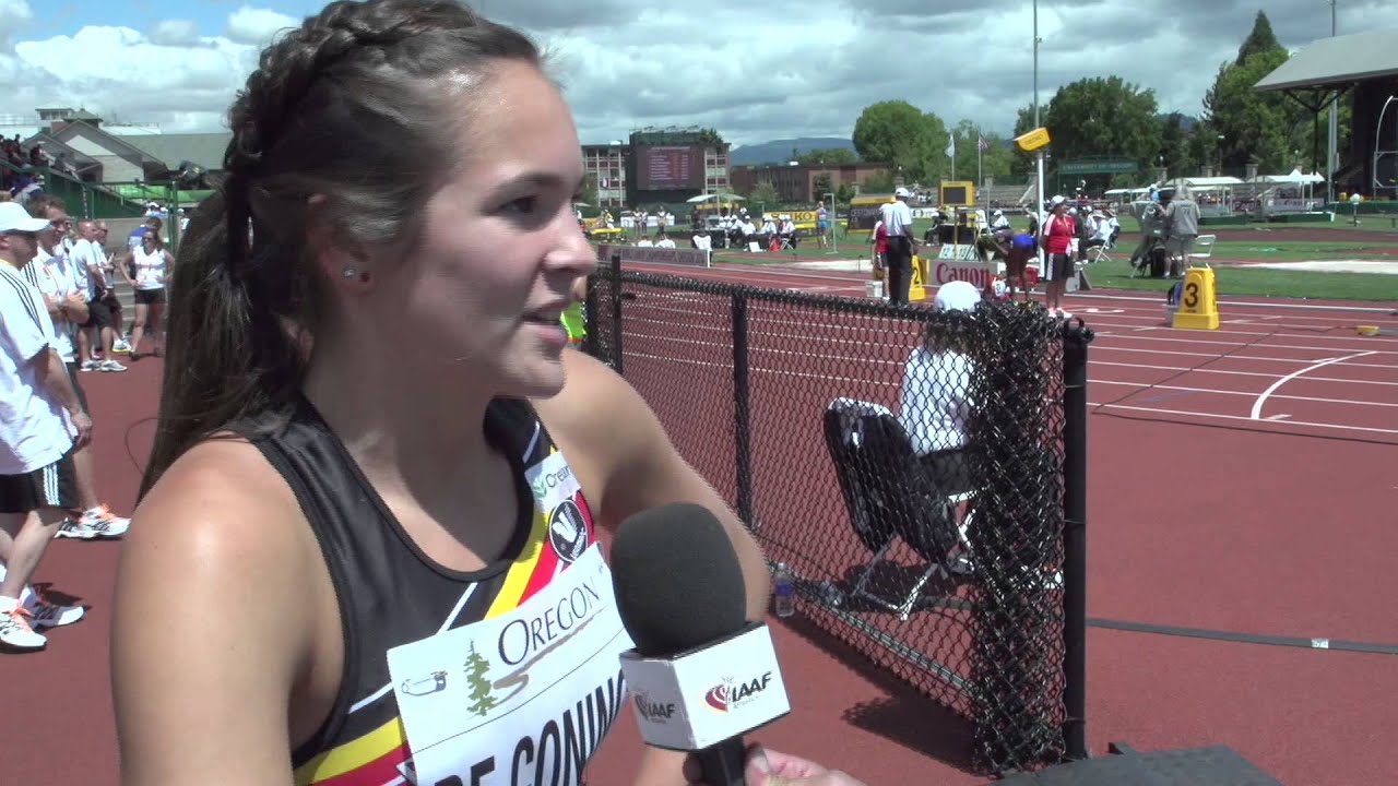 Athlete being interviewed on a track field, holding a microphone, with people and track lanes visible in the background.