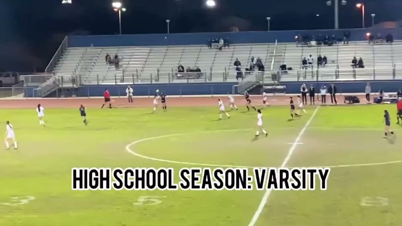 A nighttime high school soccer game in progress on a field with teams in dark and white uniforms, sparse spectators in bleachers. Text on image: "HIGH SCHOOL SEASON: VARSITY".