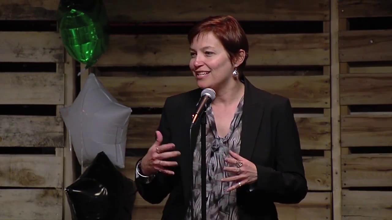 Person with short hair speaks into a microphone, gesturing with hands, standing in front of wooden pallets and star-shaped balloons.