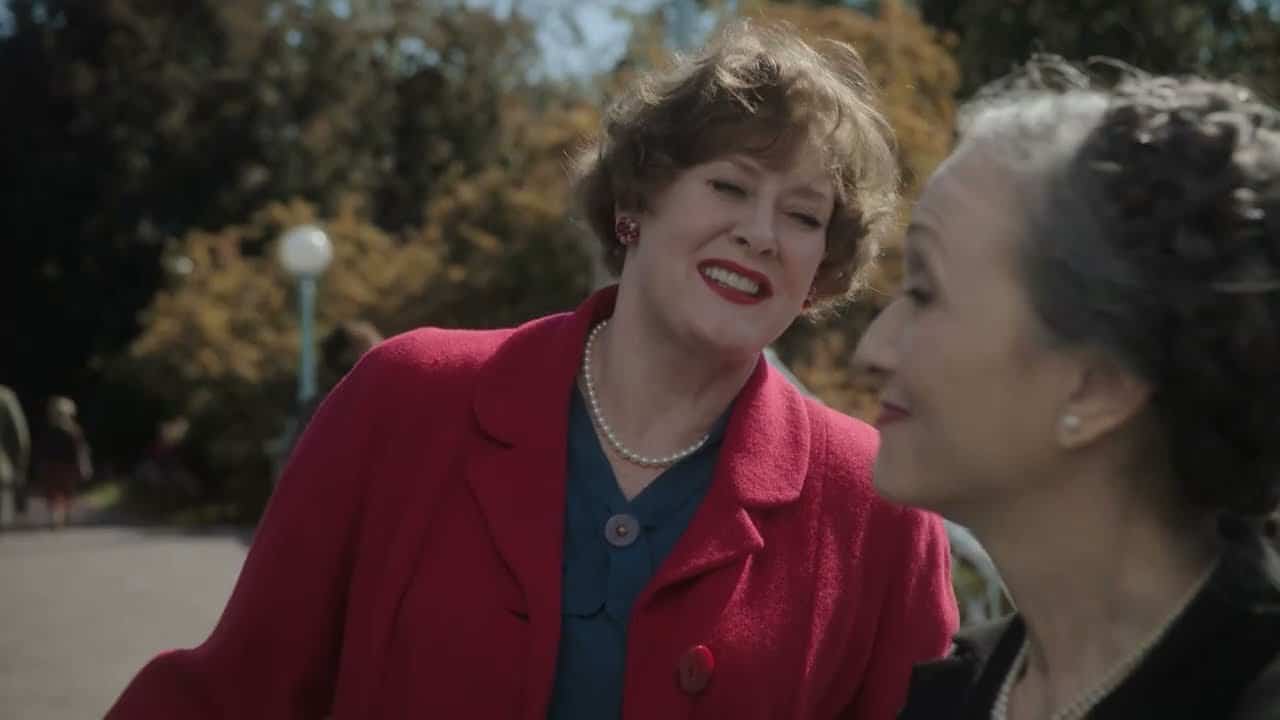 Two women are smiling and talking outdoors. One is wearing a red coat and the other has gray hair tied back. They are surrounded by trees in the background.