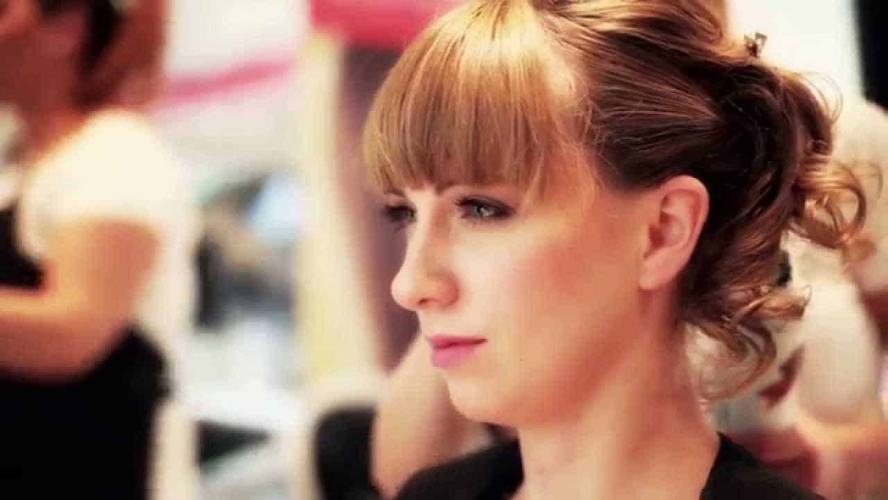 Woman with styled hair and bangs, sitting in a salon chair, blurred background showing a stylist working.