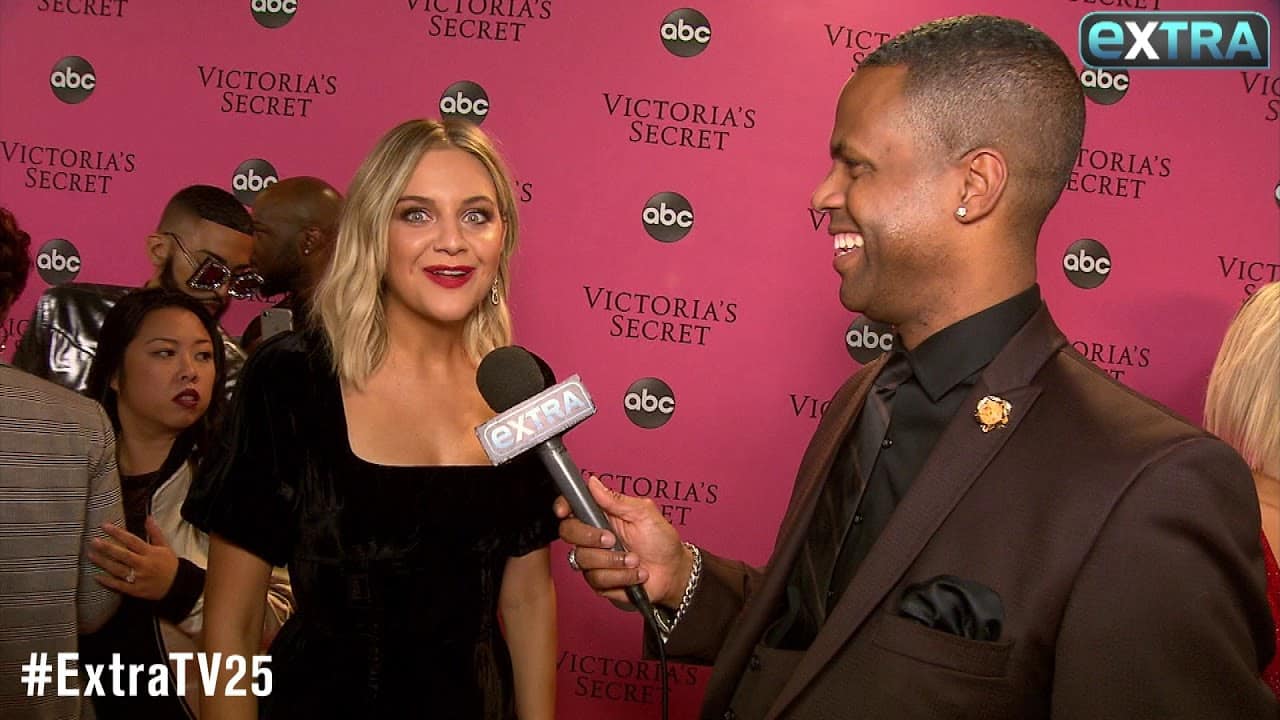 A woman in a black dress is being interviewed by a man holding an "Extra" microphone at a Victoria's Secret event with a pink backdrop.