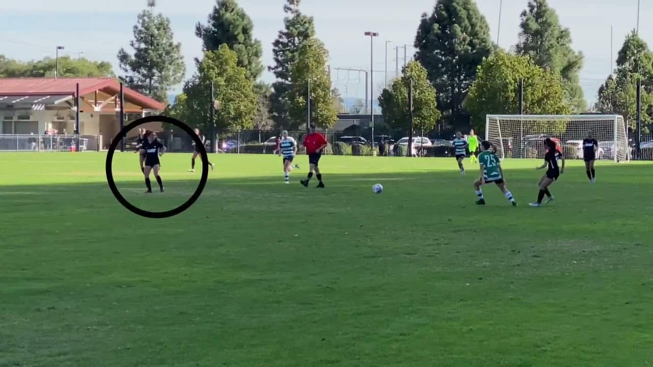 A soccer game in progress on a grassy field. Players from two teams are near the ball, with one player circled. Trees and a building are in the background.