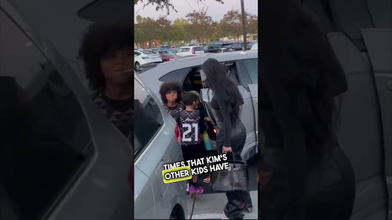 A woman stands near a minivan, holding a handbag, with two children nearby. One child wears a jersey numbered 21. Trees and parked cars are visible in the background.