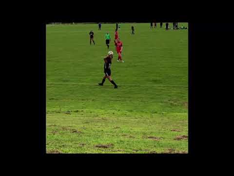 A soccer player in black walks on a grassy field, while other players and a referee are visible in the background.