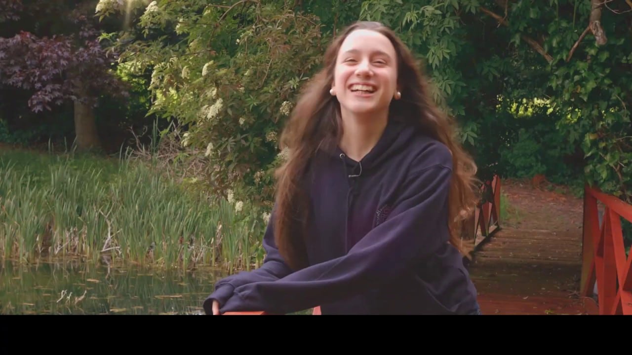 A person with long hair, wearing a dark hoodie, smiles while standing by a pond. Lush greenery is in the background.