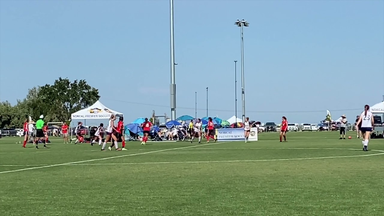 A soccer game in progress on a grassy field with players in action and spectators under tents in the background.