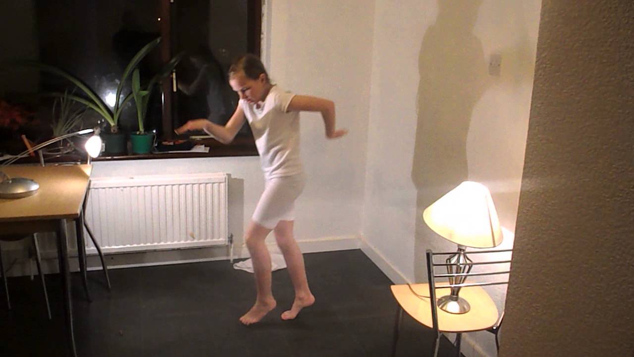 Person in a white outfit dances near a table with a lamp in a dimly lit room with potted plants on the window sill.