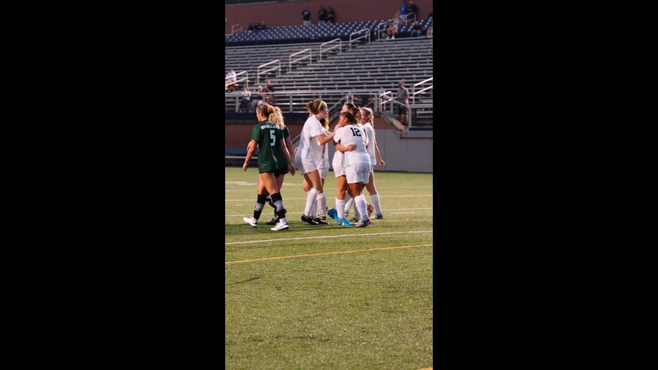 Soccer players in white uniforms celebrate on a field, while an opponent in a green uniform walks nearby. Stadium seats are visible in the background.