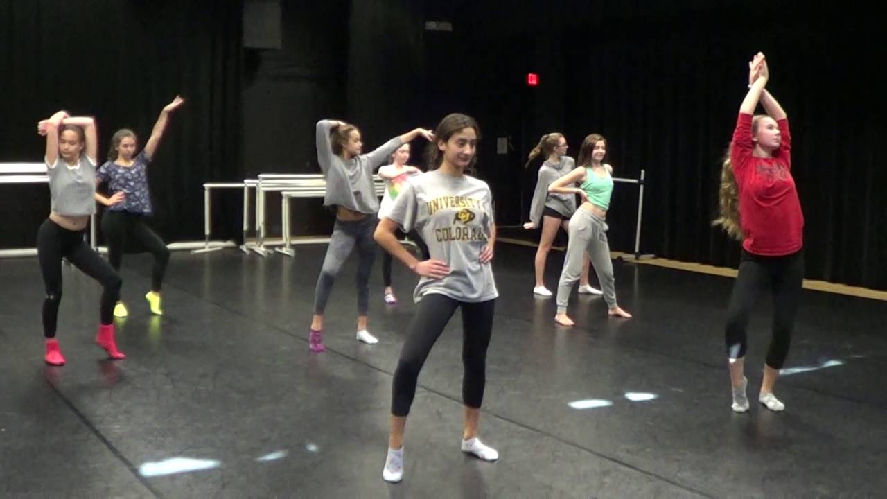 A group of young dancers rehearses in a studio, wearing casual dance attire, performing various poses and movements. The room has a black floor and walls with mirrors in the background.