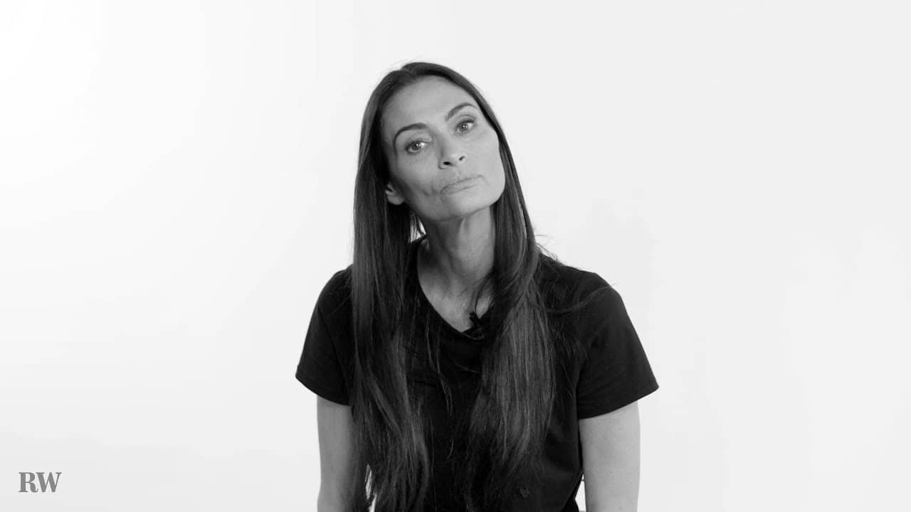 Person with long dark hair wearing a black shirt, posing against a plain white background.