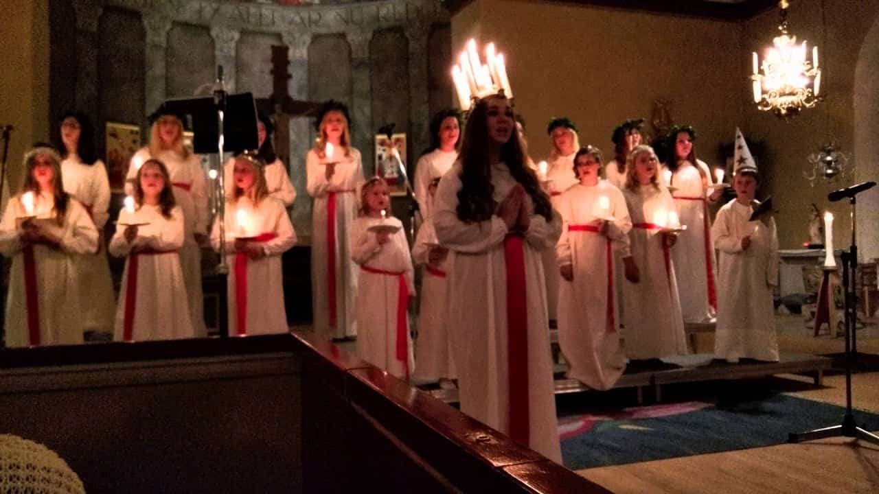 A group of people in white robes with red sashes, holding candles, perform in a dimly lit room with festive decorations.