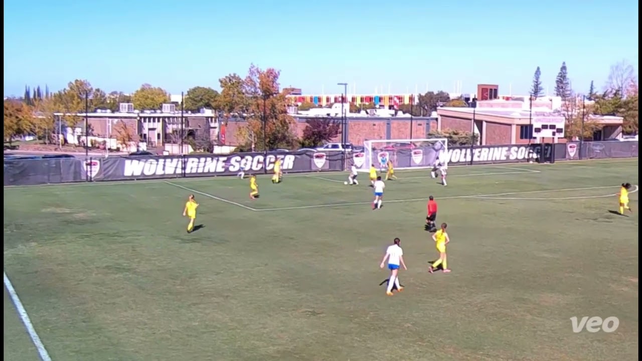 Soccer match in progress on a sunny day. Players in yellow and white uniforms on a grass field. Background shows buildings and trees.