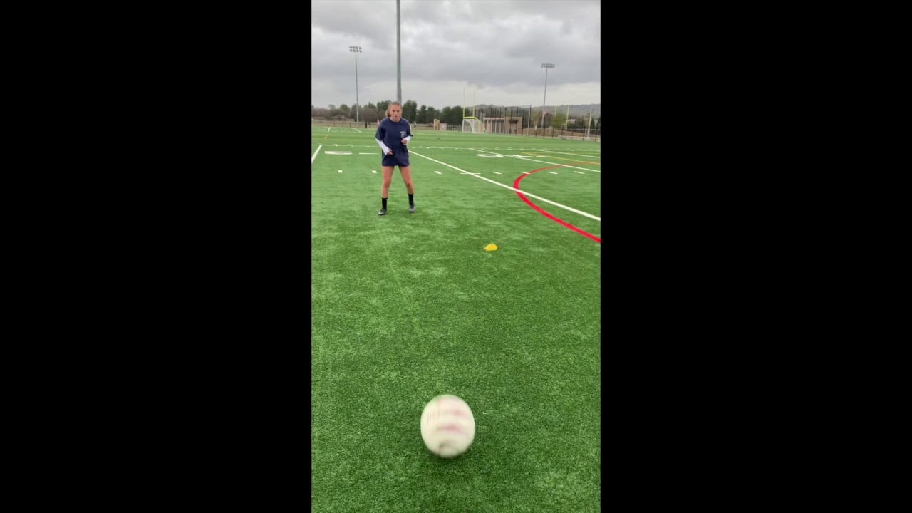 Person on a sports field in athletic gear is about to kick a ball rolling towards them. Soccer goals and cloudy sky visible in the background.