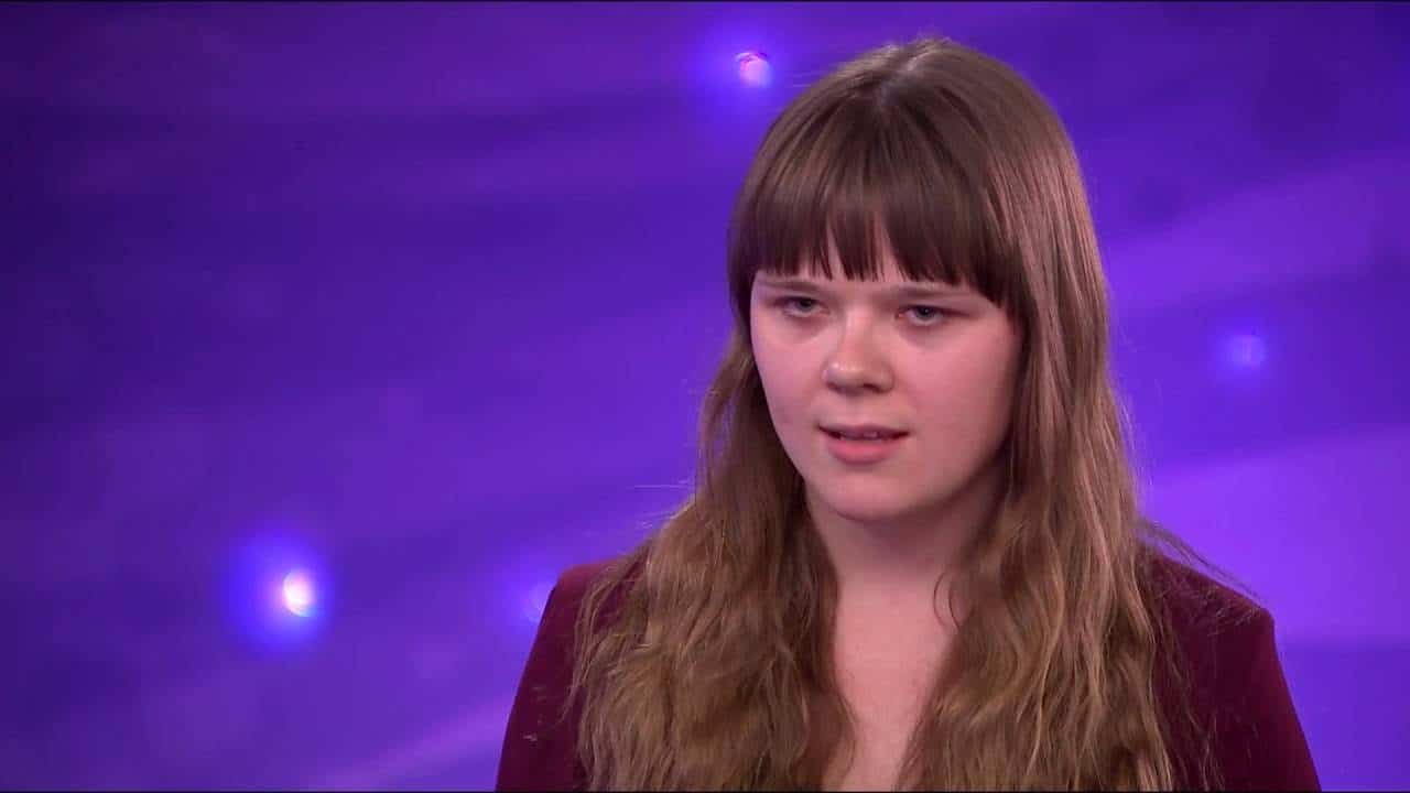 Woman with long hair and bangs stands against a purple background, looking forward.